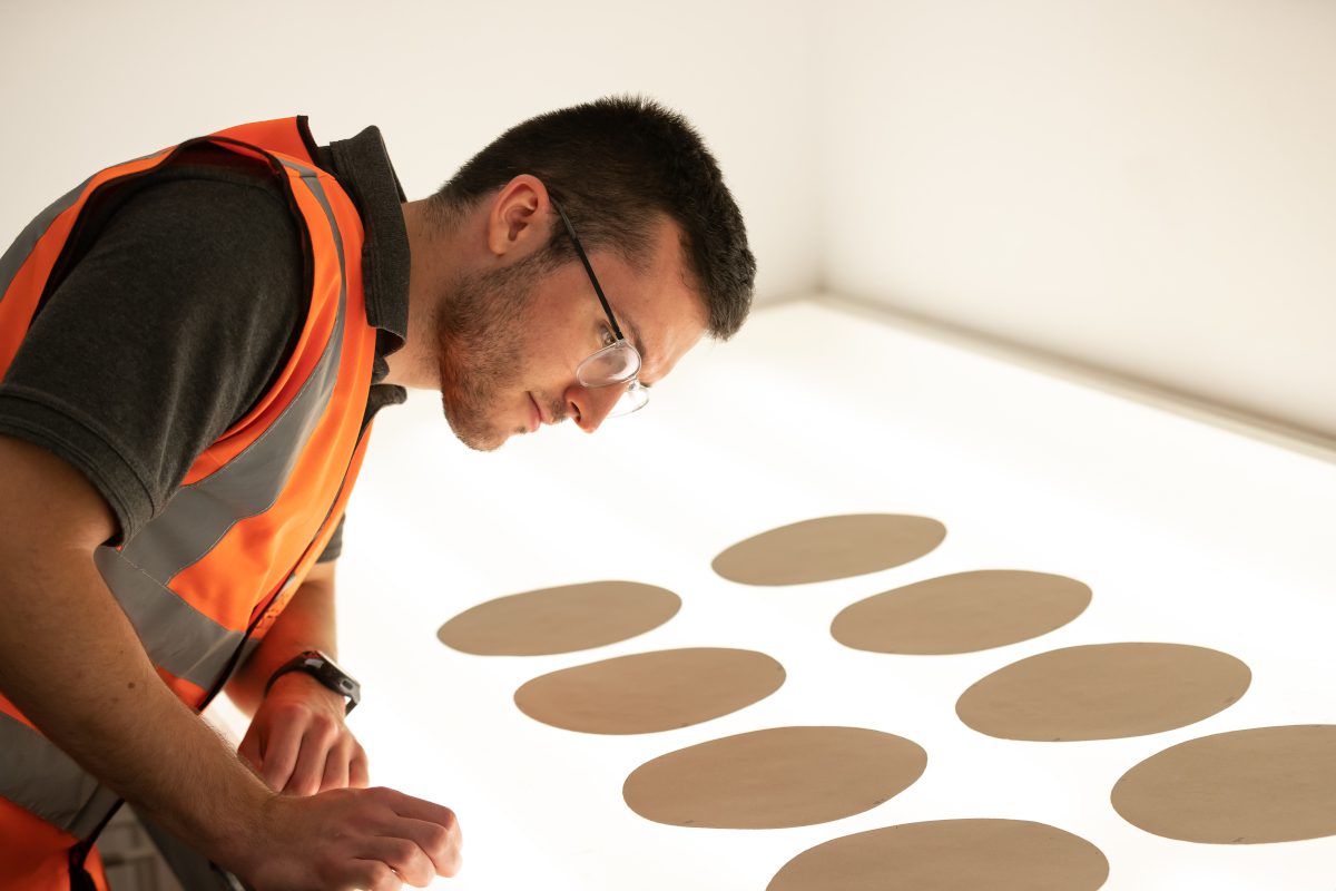 Man in luminous vest inspects sheet-like circular material lying on a lightbox or similar desk-based system, all in a professional-looking setting