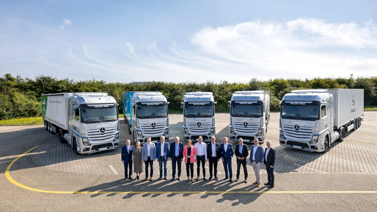 Large trucks arranged in a fan-like formation, with noses all pointing towards the camera, and a group of people standing in front of them, all in a sunny outdoor setting resembling a car park